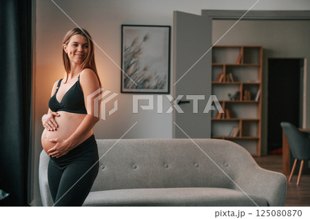 Standing and having positive mood. Beautiful pregnant woman is indoors at home Standing and having positive mood. Beautiful pregnant woman is indoors at home 125080870