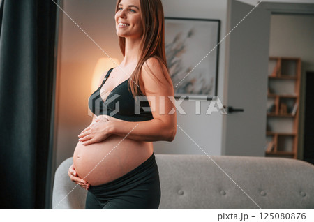 Standing near the sofa and smiling. Beautiful pregnant woman is indoors at home Standing near the sofa and smiling. Beautiful pregnant woman is indoors at home 125080876