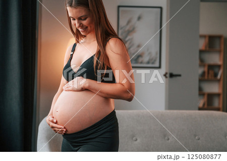 Standing near the sofa and smiling. Beautiful pregnant woman is indoors at home 125080877