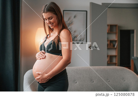 Standing near the sofa and smiling. Beautiful pregnant woman is indoors at home 125080878