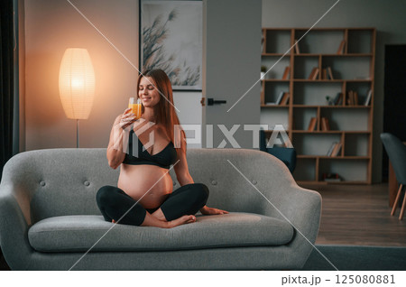 Sitting on the sofa and drinking orange juice. Beautiful pregnant woman is indoors at home 125080881