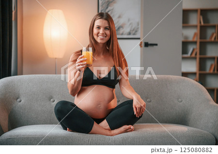 Sitting on the sofa and drinking orange juice. Beautiful pregnant woman is indoors at home Sitting on the sofa and drinking orange juice. Beautiful pregnant woman is indoors at home 125080882