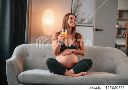 Sitting on the sofa and drinking orange juice. Beautiful pregnant woman is indoors at home 125080883