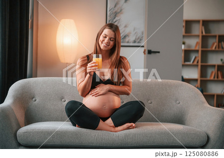With orange juice in hand. On the sofa. Beautiful pregnant woman is indoors at home 125080886