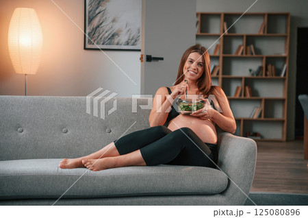 Sitting on the sofa and eating salad. Beautiful pregnant woman is indoors at home Sitting on the sofa and eating salad. Beautiful pregnant woman is indoors at home 125080896