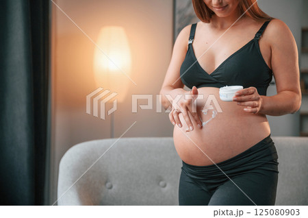 Making heart shaped picture by cream on the belly. Beautiful pregnant woman is indoors at home 125080903