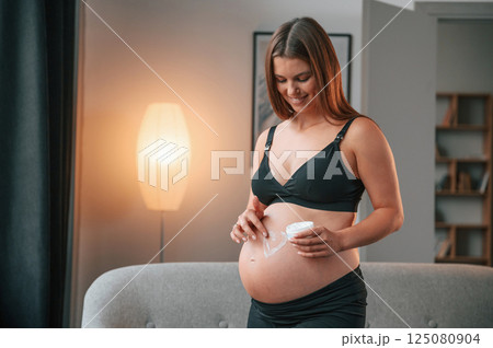 Making heart shaped picture by cream on the belly. Beautiful pregnant woman is indoors at home 125080904