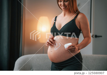 Making heart shaped picture by cream on the belly. Beautiful pregnant woman is indoors at home 125080907