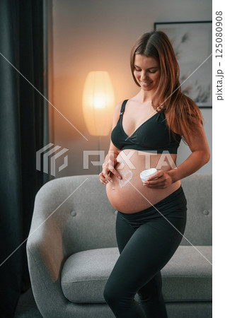 With cream on the belly. Heart shaped form. Beautiful pregnant woman is indoors at home With cream on the belly. Heart shaped form. Beautiful pregnant woman is indoors at home 125080908