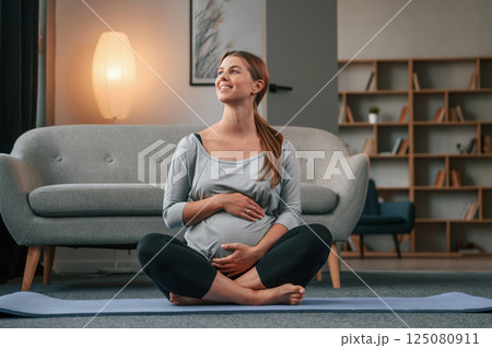 Sitting on the yoga mat. Beautiful pregnant woman is indoors at home Sitting on the yoga mat. Beautiful pregnant woman is indoors at home 125080911