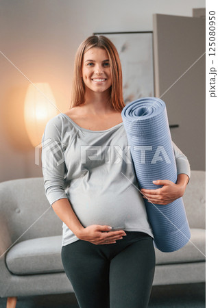 Blue colored yoga mat is in hand. Beautiful pregnant woman is indoors at home 125080950