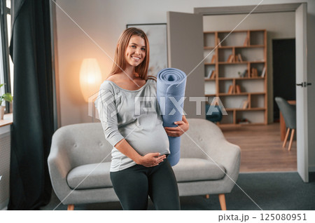 Blue colored yoga mat is in hand. Beautiful pregnant woman is indoors at home 125080951