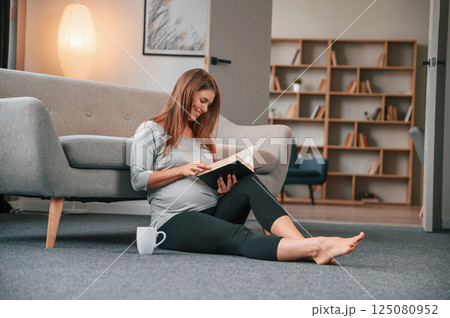 Sitting on the floor and reading the book. Beautiful pregnant woman is indoors at home Sitting on the floor and reading the book. Beautiful pregnant woman is indoors at home 125080952