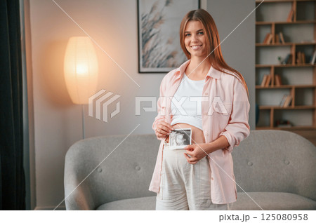 Holding ultrasound diagnostics picture of the baby. Beautiful pregnant woman is indoors at home Holding ultrasound diagnostics picture of the baby. Beautiful pregnant woman is indoors at home 125080958