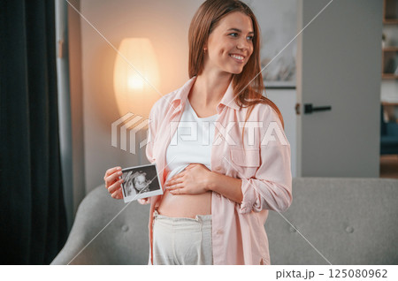 Holding ultrasound diagnostics picture of the baby. Beautiful pregnant woman is indoors at home 125080962
