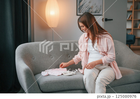 With little clothes for a baby on the sofa. Beautiful pregnant woman is indoors at home 125080976