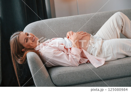 Smiling and laying down on the sofa. Beautiful pregnant woman is indoors at home Smiling and laying down on the sofa. Beautiful pregnant woman is indoors at home 125080984