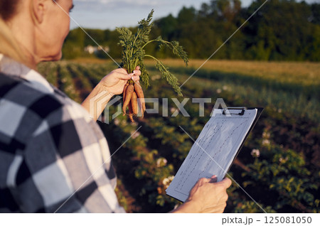 Document with information and carrots in hand. Woman is on the agricultural field at daytime 125081050