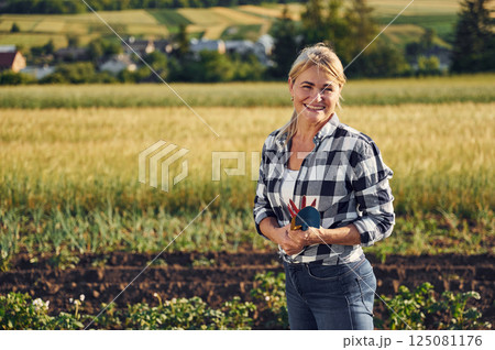 Standing and holding little showel and other garden tools. Woman is on the agricultural field at daytime 125081176