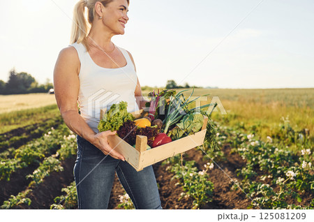Holding vegetables in wooden box. Woman is on the agricultural field at daytime 125081209