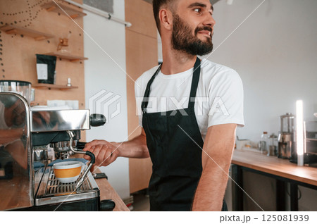 Using coffee machine. Cafe worker in white shirt and black apron is indoors Using coffee machine. Cafe worker in white shirt and black apron is indoors 125081939