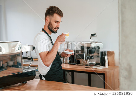 With cup of coffee. Cafe worker in white shirt and black apron is indoors 125081950