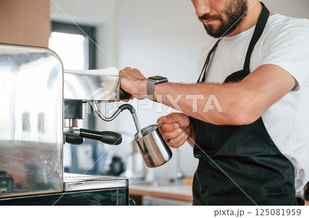 Standing and pouring coffee. Cafe worker in white shirt and black apron is indoors Standing and pouring coffee. Cafe worker in white shirt and black apron is indoors 125081959