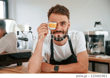 Having fun with cup. Cafe worker in white shirt and black apron is indoors Having fun with cup. Cafe worker in white shirt and black apron is indoors 125081961