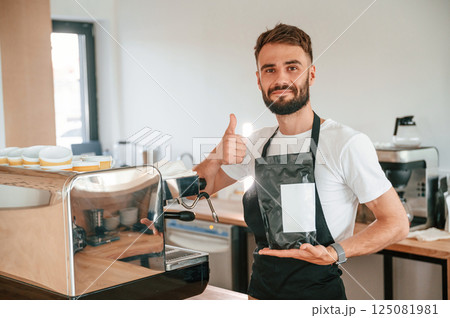 Holding package of raw coffee and showing thumb up. Cafe worker in white shirt and black apron is indoors Holding package of raw coffee and showing thumb up. Cafe worker in white shirt and black apron is indoors 125081981