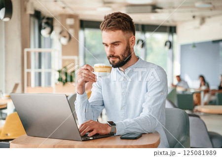 With cup of drink. Young man is sitting in the cafe and working by using laptop 125081989