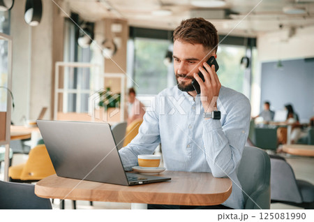 Talking by the phone. Young man is sitting in the cafe and working by using laptop 125081990