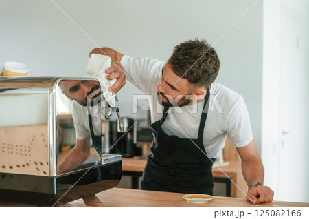Busy by cleaning coffee machine. Cafe worker in white shirt and black apron is indoors 125082166