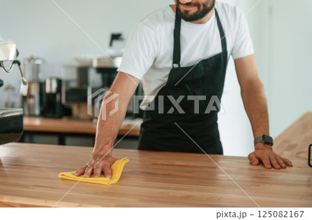 Busy by cleaning the table. Cafe worker in white shirt and black apron is indoors Busy by cleaning the table. Cafe worker in white shirt and black apron is indoors 125082167