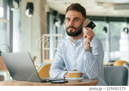 Holding credit cards. Young man is sitting in the cafe and working by using laptop Holding credit cards. Young man is sitting in the cafe and working by using laptop 125082173