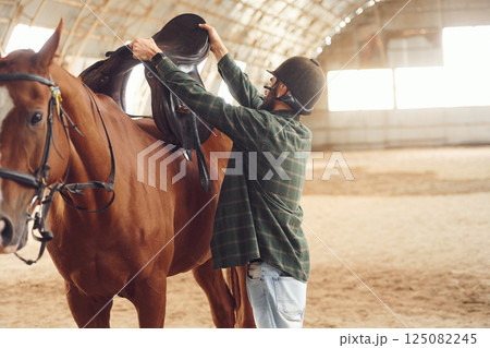 Installing the saddle. Young man with a horse is in the hangar 125082245