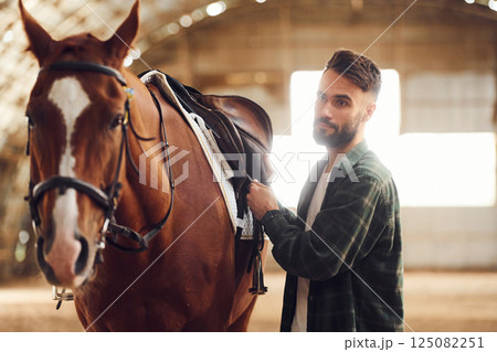 Installing the saddle. Young man with a horse is in the hangar 125082251