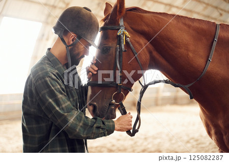 Standing and holding animal. Young man with a horse is in the hangar Standing and holding animal. Young man with a horse is in the hangar 125082287
