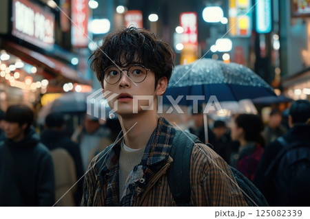 A young individual wearing glasses looks up with curiosity while navigating a bustling city street filled with bright neon signs. Rain creates a reflective surface as umbrellas pepper the crowd. 125082339