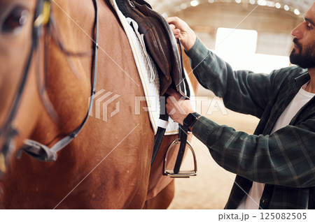 Process of preparation the animal for a ride. Young man with a horse is in the hangar 125082505