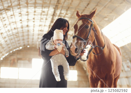 Mother with baby on the hands is standing indoors with horse in hangar 125082592