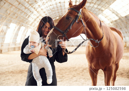 Standing and smiling. Mother with baby on the hands is indoors with horse in hangar 125082598