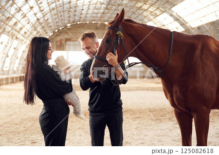 Lovely couple is standing with indoors in the hangar with little baby and showing him horse Lovely couple is standing with indoors in the hangar with little baby and showing him horse 125082618
