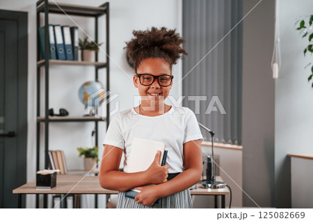 Holding paper notepad in hands. Cute african american child is standing in the domestic room Holding paper notepad in hands. Cute african american child is standing in the domestic room 125082669