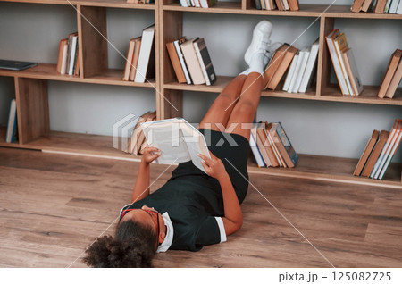 Laying down on the floor and reading the book. Cute african american girl in school uniform is at home library Laying down on the floor and reading the book. Cute african american girl in school uniform is at home library 125082725