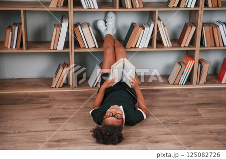 Laying down on the floor and reading the book. Cute african american girl in school uniform is at home library Laying down on the floor and reading the book. Cute african american girl in school uniform is at home library 125082726