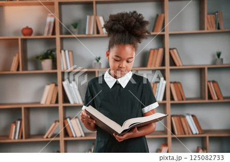 Standing and reading the book. Cute african american girl in school uniform is at home library 125082733