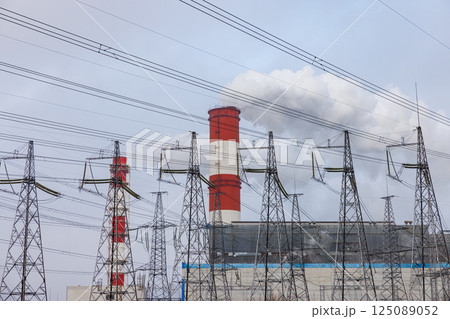 Large chimneys release white smoke in an industrial landscape filled with electricity pylons under a gray sky 125089052