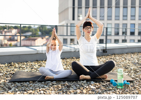 Mother and young daughter sitting on rooftop practicing meditation and yoga in outdoor environment. Family bonding through fitness and healthy living. Wearing casual sports clothing Mother and young daughter sitting on rooftop practicing meditation and yoga in outdoor environment. Family bonding through fitness and healthy living. Wearing casual sports clothing 125089707