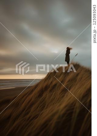 Lone figure on beach dune facing windy sunset sky at seaside 125093043