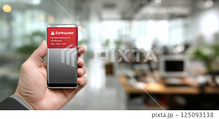 hand holding smartphone with earthquake alert displayed on screen, set in blurred office hand holding smartphone with earthquake alert displayed on screen, set in blurred office 125093138